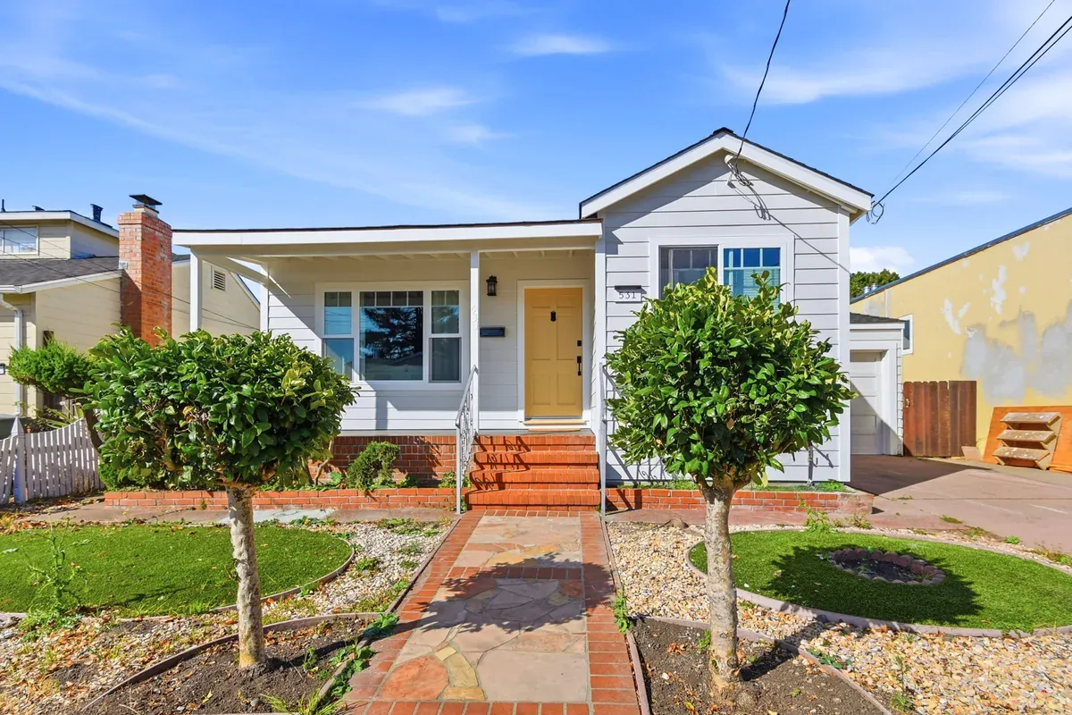 Front of the white Craftsman bungalow with a yellow door, topiary trees and a brick-edged flagstone walk