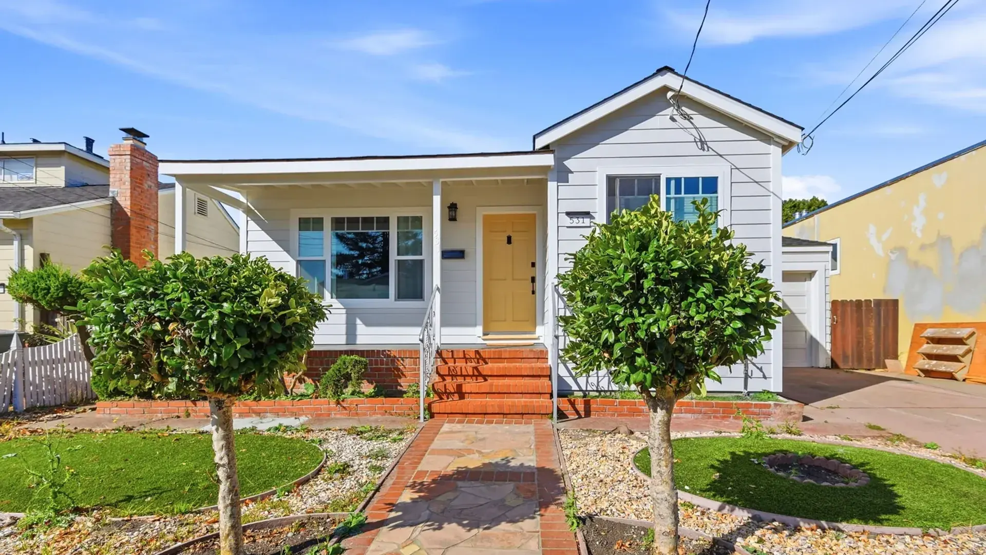 Front of a white single-story bungalow with a covered porch, yellow front door, and brick steps, framed by two topiary trees along a flagstone walkway